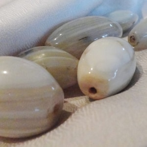 May include: A collection of six smooth, white agate stones with brown and gray banding. The stones are arranged on a white fabric background.