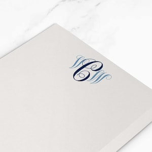 Puede incluir: Una pila de blocs de notas blancos con un monograma azul marino en la esquina superior derecha. El bloc de notas mide 10,8 x 15,9 cm. Dos clips plateados en forma de estrella están en el fondo.