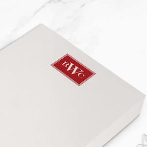 May include: A white notepad with a red rectangular logo in the upper right corner. The logo contains the letters "BWC". The notepad measures approximately 10.8 x 15.9 cm. Two silver star-shaped paper clips are in the background.