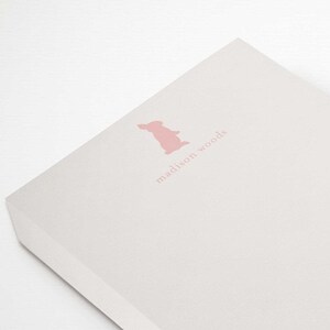 Puede incluir: Un bloc de notas blanco con un logotipo de conejo rosa y el texto "madison woods". Un lápiz rosa se sostiene sobre el bloc de notas. El bloc de notas mide aproximadamente 12,7 x 17,8 cm.