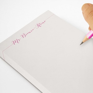 Puede incluir: Un bloc de notas blanco con el texto "Ms. Bianca Kline" en escritura fucsia. Un lápiz rosa se sostiene sobre el bloc de notas. El bloc de notas mide aproximadamente 12,7 x 17,8 cm.
