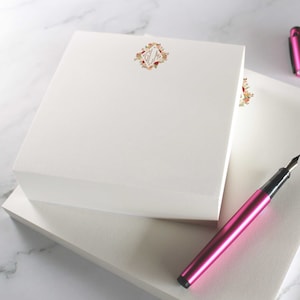 May include: White notepads with a floral monogram, a pink fountain pen, and an inkwell. The notepads are square and textured. The pen is a bright pink colour. The inkwell is black.