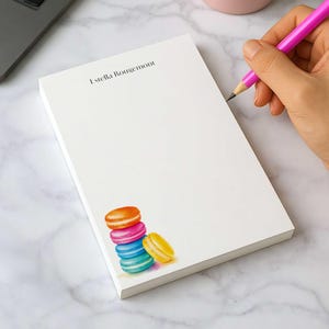 May include: A white rectangular notepad featuring the printed name Estella Rougemont at the top and a colorful illustration of a stack of four macarons in orange, pink, blue, and yellow at the bottom corner, resting on a marble surface.