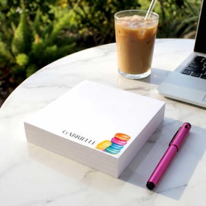 May include: A square white notepad featuring the name Gabrielle printed in black text next to a small illustration of four colorful stacked macarons in yellow, blue, pink, and orange. The notepad is positioned on a white surface.