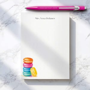 May include: A white rectangular notepad featuring the printed name Mrs. Vera Delaney at the top and a colorful illustration of a stack of four macarons in orange, pink, blue, and yellow at the bottom, accompanied by a bright pink ballpoint pen.