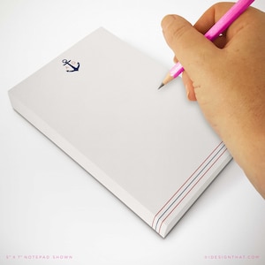 Puede incluir: Bloc de notas blanco con un diseño de ancla azul marino y rayas rojas y azules. El bloc de notas mide 12,7 x 17,8 cm. Un lápiz rosa se sostiene sobre las páginas en blanco, listo para escribir.