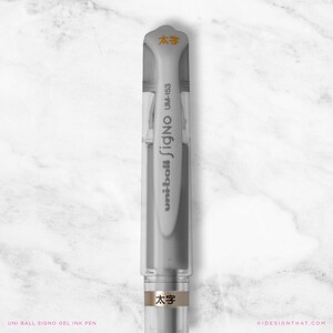 May include: A clear and grey Uni-ball Signo gel ink pen, model UM-153, with the text "太字" in orange. The pen has a clear cap and a grey body. The pen is on a white marble surface.