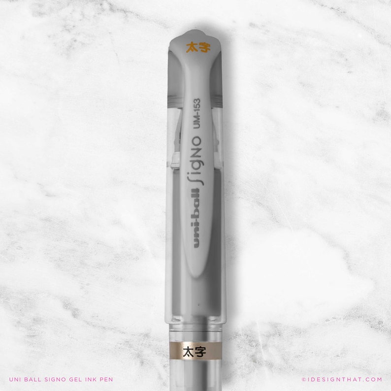 May include: A clear and grey Uni-ball Signo gel ink pen, model UM-153, with the text "太字" in orange. The pen has a clear cap and a grey body. The pen is on a white marble surface.