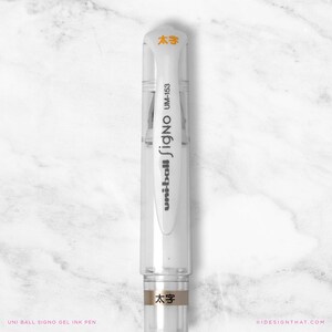 May include: A clear Uni-ball Signo gel ink pen with white accents and orange text. The pen has the text "uni-ball Signo UM-153" printed on the side. The pen also has the Japanese characters "太字" printed on the top and bottom.