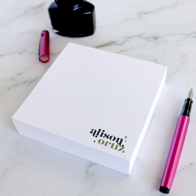 Op de afbeelding: Een wit notitieblok met de tekst "alison.ortiz." gedrukt in zwart en groen in de rechterbenedenhoek. Het notitieblok ligt op een wit marmeren oppervlak met een roze pen en een zwarte inktpot.