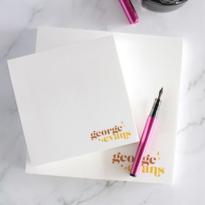 Op de afbeelding: Twee witte notitieblokken met de tekst "george.evans" gedrukt in bruine en gouden letters. Een roze vulpen ligt op de notitieblokken.