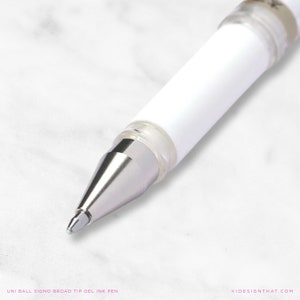 May include: Close-up of a white Uni Ball Signo broad tip gel ink pen. The pen has a silver tip and a clear plastic section near the tip. The pen's body is white. The text "UNI BALL SIGNO BROAD TIP GEL INK PEN" is at the bottom.
