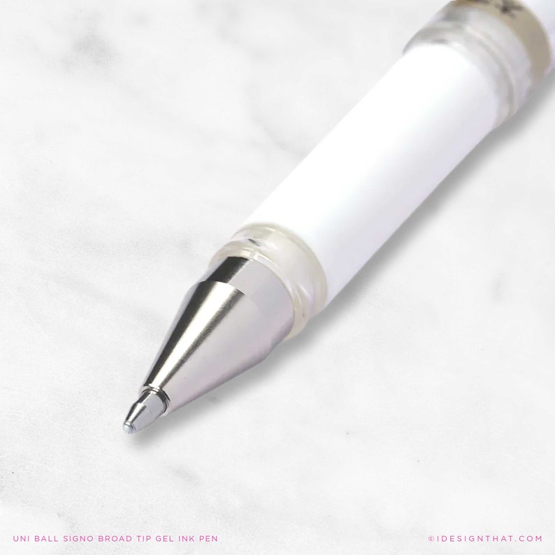 May include: Close-up of a white Uni Ball Signo broad tip gel ink pen. The pen has a silver tip and a clear plastic section near the tip. The pen's body is white. The text "UNI BALL SIGNO BROAD TIP GEL INK PEN" is at the bottom.