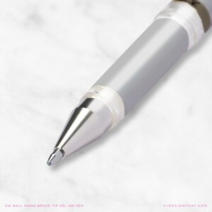 May include: A close-up shot of a silver and clear Uni-ball Signo Broad Tip gel ink pen. The pen features a silver barrel and a transparent grip. The pen's tip is in focus, with the text "UNI BALL SIGNO BROAD TIP GEL INK PEN" at the bottom.
