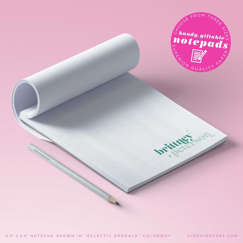 Op de afbeelding: Een wit notitieboekje met de tekst "brittney peterson" in het groen gedrukt op de voorkant. Het notitieboekje is 14 cm bij 14 cm en wordt getoond in de kleur "eclectische smaragd". Een grijs potlood ligt naast het notitieboekje.