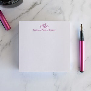 Puede incluir: Un bloc de notas blanco con un gráfico de bicicleta magenta y el texto "Gemma Pearl Bailey". Una pluma estilográfica magenta y un portalápices sobre una superficie de mármol.