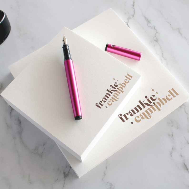 Op de afbeelding: Twee witte notitieblokken met de tekst "frankie, campbell" in bruin gedrukt op de voorkant. Een roze vulpen en een roze markeerstift liggen op de notitieblokken.