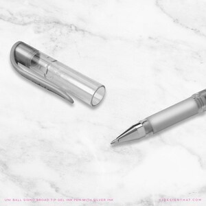 May include: A silver and clear gel ink pen with the cap removed, on a white marble surface. The pen's tip is visible, and the text "UNI BALL SIGNO BROAD TIP GEL INK PEN WITH SILVER INK" is at the bottom.