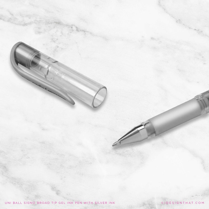 May include: A silver and clear gel ink pen with the cap removed, on a white marble surface. The pen's tip is visible, and the text "UNI BALL SIGNO BROAD TIP GEL INK PEN WITH SILVER INK" is at the bottom.