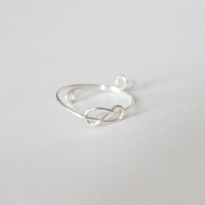 K&ouml;nnte beinhalten: Ein zarter Silberring mit einem Knoten-Design. Der Ring ist aus d&uuml;nnem, gl&auml;nzendem Metall gefertigt und hat ein schlichtes, elegantes Aussehen. Der Knoten ist der Mittelpunkt und verleiht dem Schmuck einen Hauch von einzigartigem Stil.