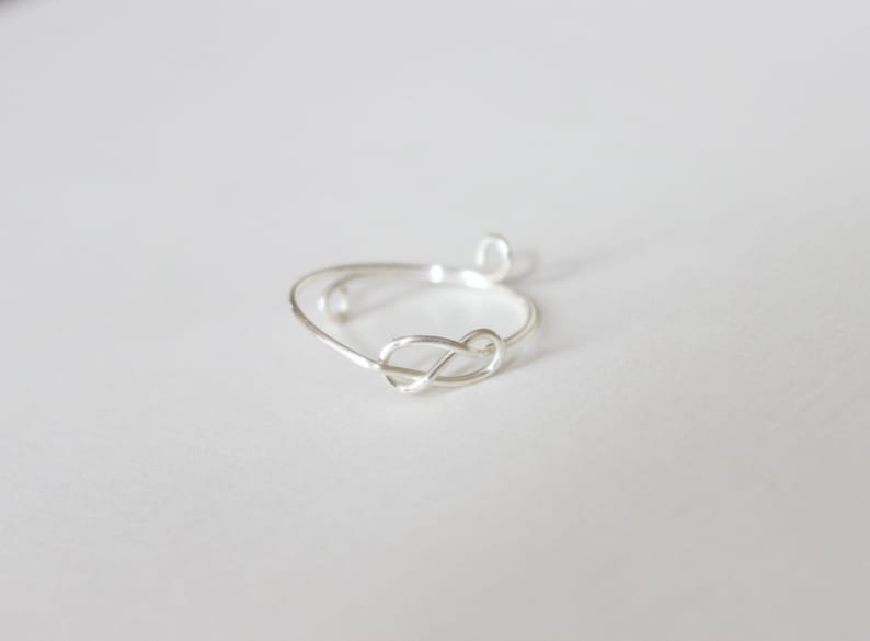 K&ouml;nnte beinhalten: Ein zarter Silberring mit einem Knoten-Design. Der Ring ist aus d&uuml;nnem, gl&auml;nzendem Metall gefertigt und hat ein schlichtes, elegantes Aussehen. Der Knoten ist der Mittelpunkt und verleiht dem Schmuck einen Hauch von einzigartigem Stil.