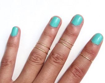 May include: A hand wearing multiple thin gold rings on the middle and ring fingers. The fingernails are painted a light turquoise color.