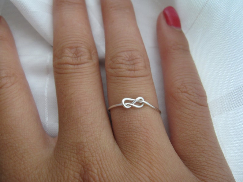 K&ouml;nnte beinhalten: Ein silberner Ring mit einem Knoten-Design. Der Ring ist d&uuml;nn und filigran, wobei der Knoten im Mittelpunkt steht. Der Ring wird an einem Finger getragen und zeigt seine schlichte Eleganz.