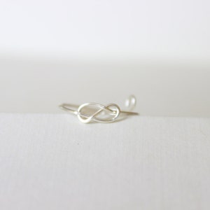 K&ouml;nnte beinhalten: Ein zarter Silberring im Knoten-Design. Der Ring besteht aus d&uuml;nnem, gl&auml;nzendem Metall und liegt auf einer wei&szlig;en Oberfl&auml;che. Der Knoten ist der Mittelpunkt, mit einem kleinen, runden Detail am Ende des Bandes.
