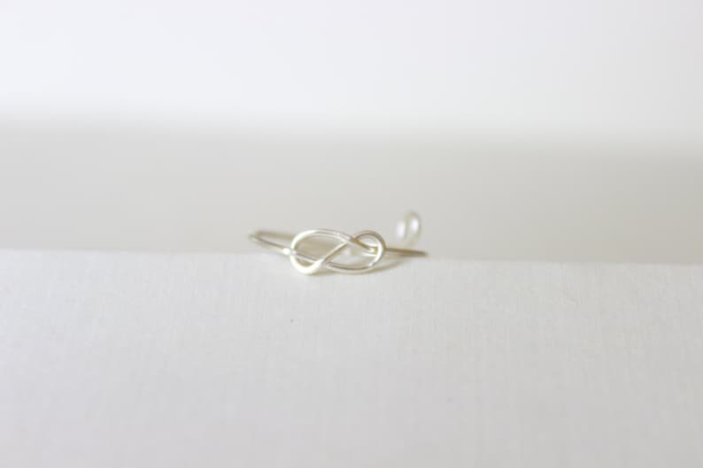 K&ouml;nnte beinhalten: Ein zarter Silberring im Knoten-Design. Der Ring besteht aus d&uuml;nnem, gl&auml;nzendem Metall und liegt auf einer wei&szlig;en Oberfl&auml;che. Der Knoten ist der Mittelpunkt, mit einem kleinen, runden Detail am Ende des Bandes.