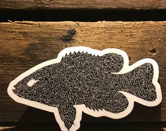 May include: A black and white fish sticker with a detailed design made up of small, repeating shapes. The sticker is on a wooden background.