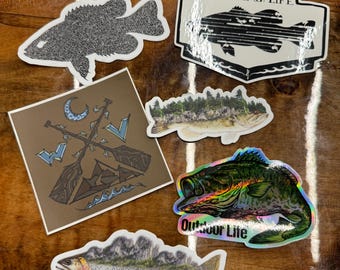 May include: Assortment of six fishing-themed stickers. The stickers feature various fish designs, including a black and white bass, a colorful bass, a trout, and a fish with a forest scene. The stickers also include a design with two paddles and a crescent moon and a sticker with the text "Mountain Life" and a black and white bass.