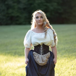 May include: A woman wearing a white peasant top with a black corset and a long blue skirt. She is standing in a field of green grass.