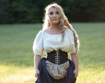 May include: A woman wearing a white peasant top with a black corset and a long blue skirt. She is standing in a field of green grass.