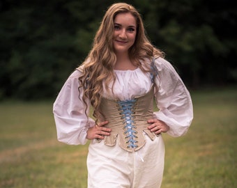 May include: A woman wearing a white blouse with a beige corset and white pants. The corset has blue ribbon lacing and is tied at the front. She is standing in a grassy field.