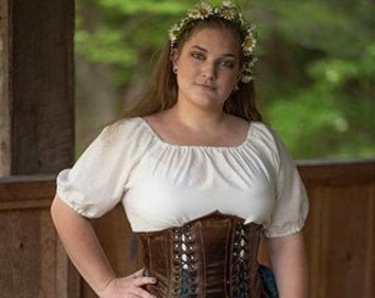 May include: A woman wearing a white peasant blouse, a brown leather corset with lacing, and a green and blue plaid skirt. She is wearing a flower crown.