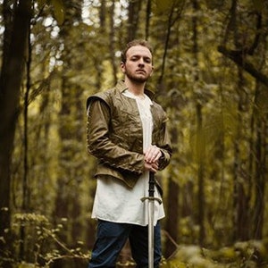 May include: A man in a forest setting, wearing a brown leather jacket and white shirt, holds a silver sword in front of him.