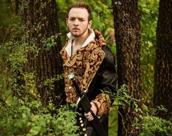 May include: A man in a forest setting, wearing a black coat with a gold and red patterned lining. He is holding a sword in his right hand.