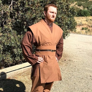 May include: A person wearing a brown robe with a brown belt and brown boots. The robe has a long, flowing skirt and a collar that is folded down. The person is standing in a dirt lot with trees in the background.