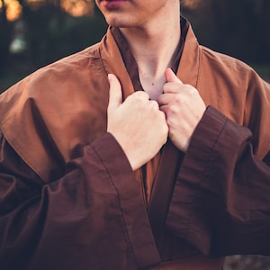 May include: A person wearing a brown robe with a belt. The robe has a collar that is being held closed by the person's hands.