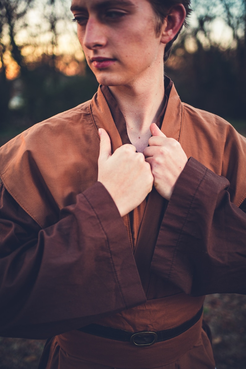 May include: A person wearing a brown robe with a belt. The robe has a collar that is being held closed by the person's hands.