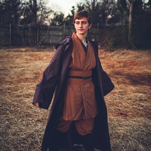 May include: A person wearing a brown and purple robe with a belt. The person is standing in a field of dry grass.