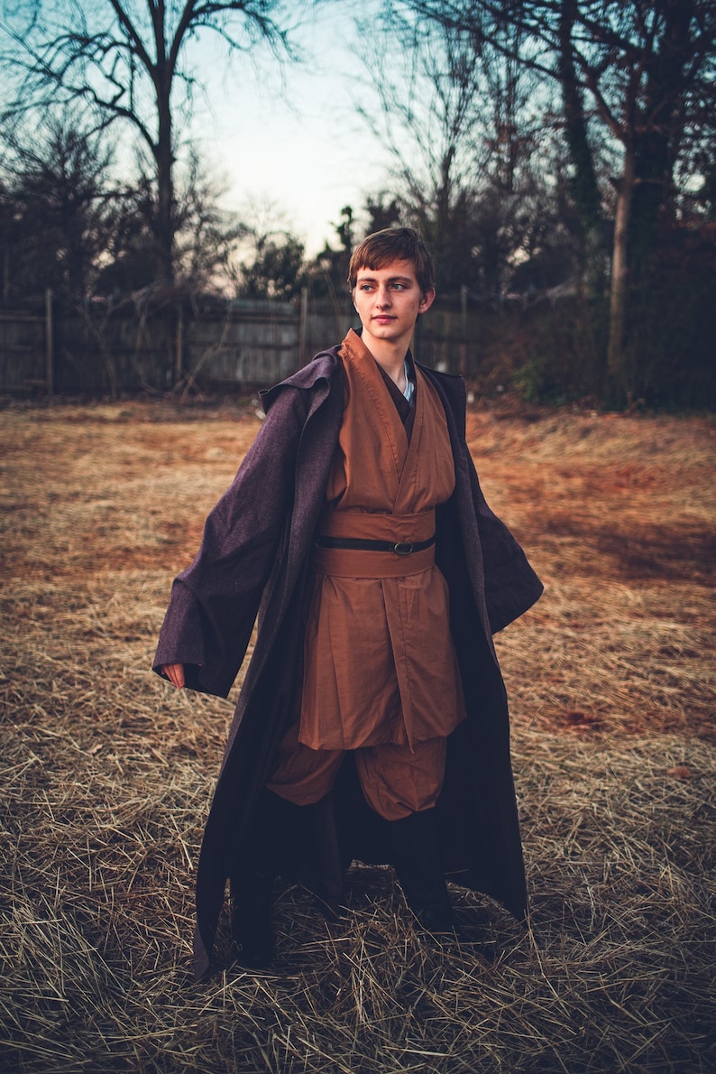 May include: A person wearing a brown and purple robe with a belt. The person is standing in a field of dry grass.