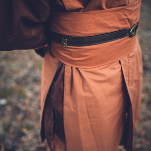 May include: A long-sleeved garment in brown, featuring a brown leather belt with a metal buckle. The garment has a gathered waist and a flowing skirt.