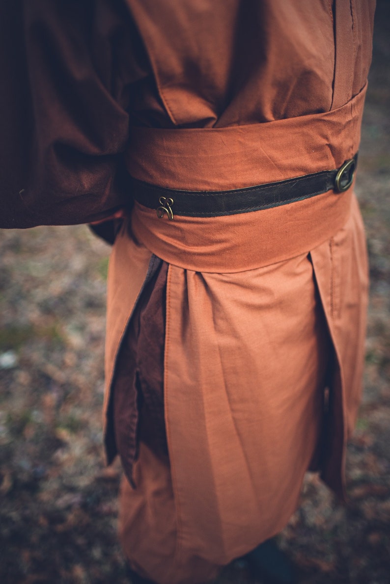 May include: A long-sleeved garment in brown, featuring a brown leather belt with a metal buckle. The garment has a gathered waist and a flowing skirt.