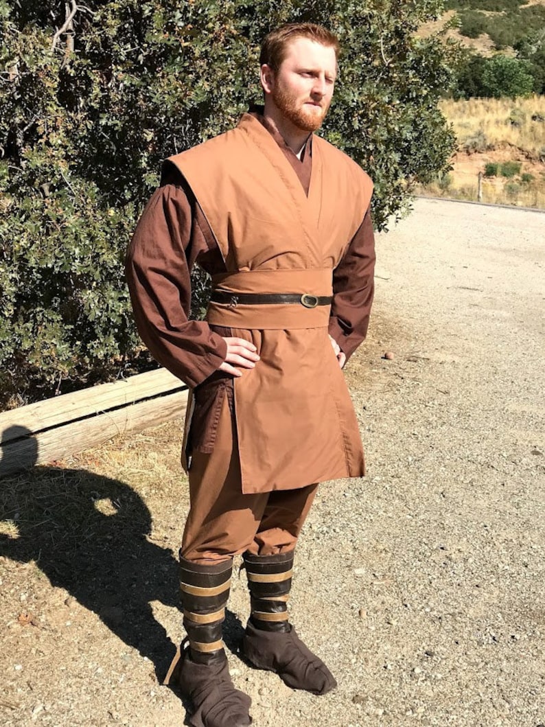 May include: A person wearing a brown robe with a brown belt and brown boots. The robe has a long, flowing skirt and a collar that is folded down. The person is standing in a dirt lot with trees in the background.
