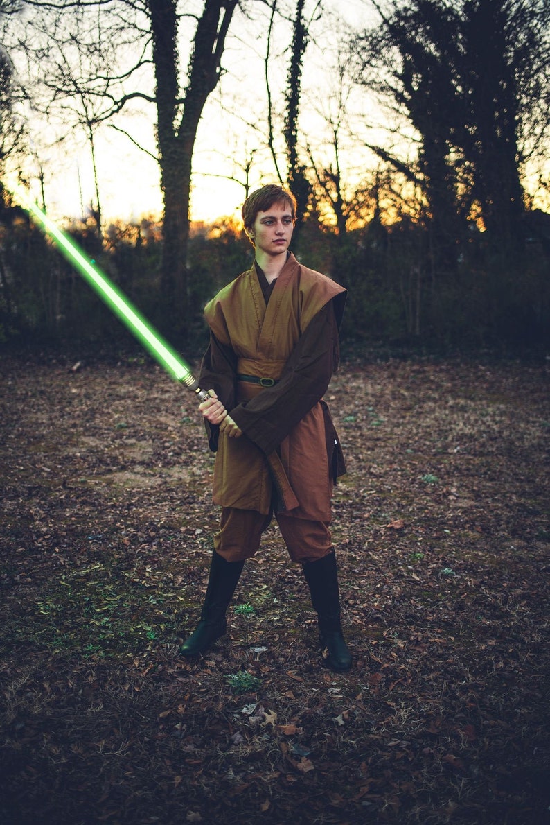May include: A person wearing a brown robe holds a green lightsaber in a woodland setting.