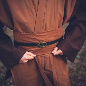 May include: A brown robe with a black belt and a metal buckle. The robe is being worn by a person, but the person is not visible in the image.