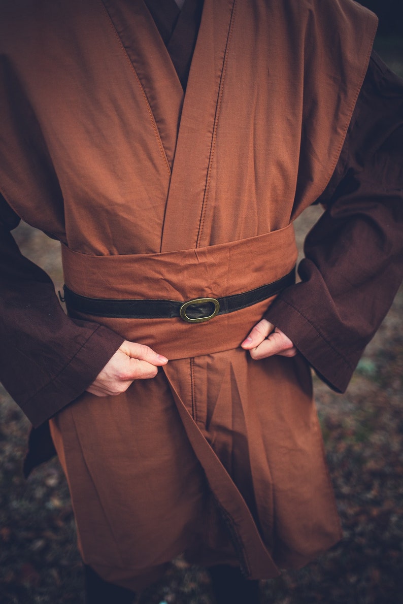 May include: A brown robe with a black belt and a metal buckle. The robe is being worn by a person, but the person is not visible in the image.