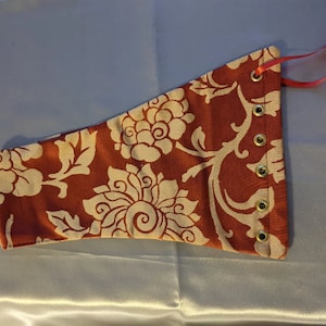 May include: A red fabric cuff with a white floral pattern and gold eyelets. The cuff is tied with a red ribbon.