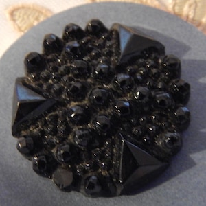 May include: A round, gray button with a black, ornate design. The button's center features a cluster of small, round beads and four larger, triangular shapes. The button is set against a cream-colored background with a floral lace pattern.